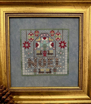 A framed needlepoint artwork featuring a house with birds, floral motifs, and the word "NOR" stitched at the bottom, set in a blue-gray fabric and enclosed in an ornate gold frame.
