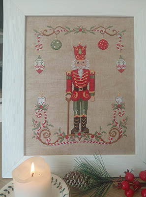 Cross stitch design featuring a red and green nutcracker surrounded by ornaments, greenery, and a decorative holiday border on a beige background.