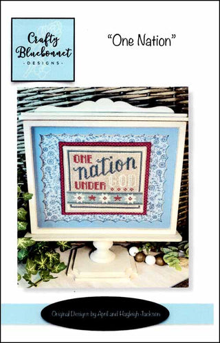 Patriotic cross stitch pattern featuring the phrase “One Nation Under God” in red, white, and blue tones with vintage Americana accents, framed for display, by Crafty Bluebonnet Designs.
