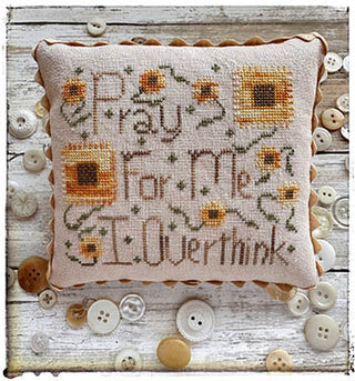 Finished cross-stitch pillow with the phrase ‘Pray For Me, I Overthink’ surrounded by golden sunflowers on neutral fabric, designed by Lucy Beam.