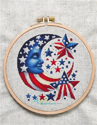 Embroidery hoop with a patriotic cross stitched design featuring stars, stripes, and a moon on a textured fabric background.