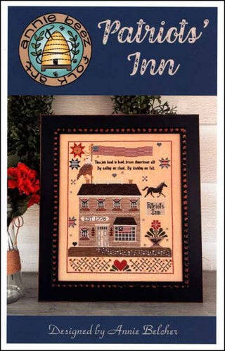 Framed cross-stitch pattern of a house with decorative elements, featuring the Annie Beez logo.