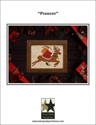 Framed cross-stitched picture of a reindeer and Santa on a wooden surface with Christmas decorations.