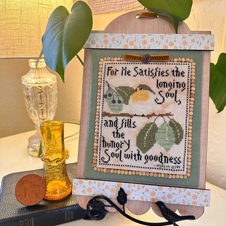 Decorative cross-stitched piece with birds and text on a stand, surrounded by plants and a lamp.