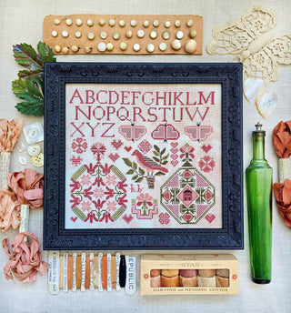 Framed cross-stitch sampler featuring a red alphabet across the top and multiple Quaker-style heart motifs in red and green geometric patterns on neutral fabric with embroidery tools and materials on a textured surface