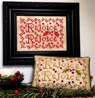 Framed cross-stitched piece with 'Rejoice' text and a matching pillow on a decorative background.