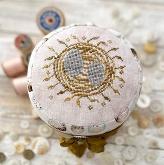 Finished drum-style pincushion topped with a cross-stitched nest containing two blue robin eggs, surrounded by warm brown straw motifs on pale linen, designed by Lucy Beam.