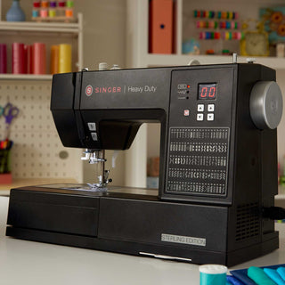 Singer heavy-duty sewing machine on a table with a blurred background of colorful spools of thread and sewing supplies.