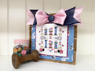 Decorative spool with a pink and blue bow, jar of buttons, and cross-stitch pattern on a white background