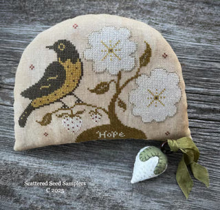 Cross stitch model for Seeds of Hope Pinkeep & Strawberry by Scattered Seed Samplers featuring a black and golden-brown bird, large white flowers, berries and the word Hope, finished as a primitive pinkeep with a coordinating white strawberry charm.