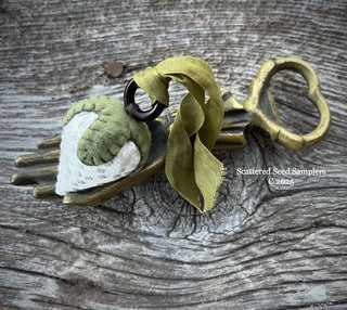 A vintage-style metal key-shaped seed sampler charm with a green and white fabric bird, olive green ribbon, and brass ring, displayed on a weathered gray wooden surface. A watermark reads 'Scattered Seed Samplers' with a copyright notice.