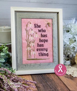 Framed cross stitch sampler on soft pink fabric featuring the phrase “She who has hope has every thing” with delicate floral motifs and a small bee accent.
