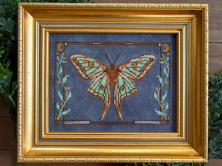 Cross stitch pattern featuring a mint-green moth with brown and gold detailing, surrounded by crescent moons and leafy accents on a denim blue background