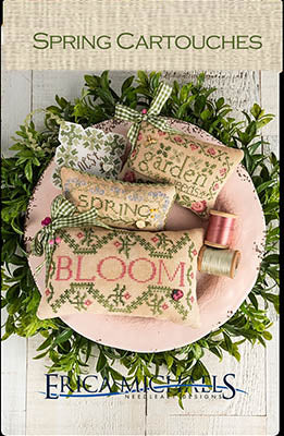 Spring-themed cross-stitch cartouches on a wooden surface with greenery