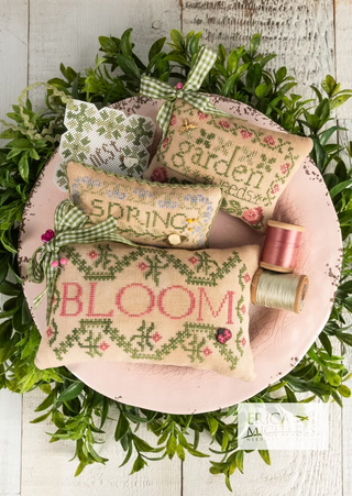 Decorative pillows with cross-stitch patterns on a wooden plate surrounded by greenery