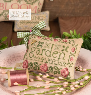 Decorative pillow with cross-stitch design and pink roses, surrounded by plants and a spool of thread, with Erica Michaels embroidery studio branding.