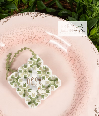 Cross-stitched ornament with 'nest' on a pink textured surface, brand 'Erica Michiels' visible.