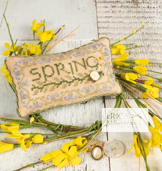 Decorative pillow with 'Spring' embroidery surrounded by yellow flowers on a wooden surface.