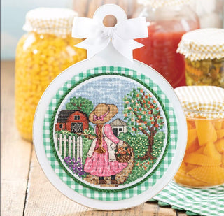 Decorative cross-stitched ornament with a woman in a garden and barn, surrounded by jars of canned goods.