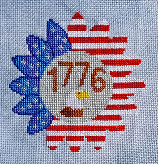 Patriotic sunflower cross stitch design featuring red and white striped petals, blue background with white stars, the number “1776” in red above a stitched eagle, set against a tan fabric.
