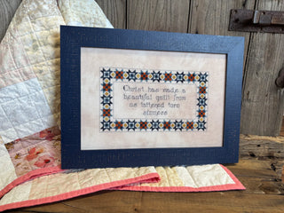 Framed cross-stitched sampler with decorative border and text on a wooden surface with quilt in the background.