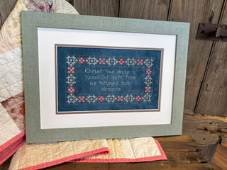 Cross stitch sampler featuring a religious quote about Christ making a quilt from tattered sinners, framed by a geometric quilt-style border in red, blue, and gold tones on neutral fabric