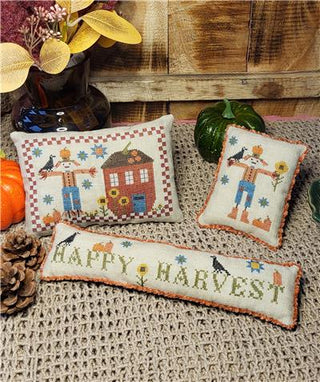 Three small cross-stitched pillows with scarecrows, pumpkins, sunflowers, and Happy Harvest text in warm fall colors on beige fabric.