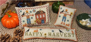 Three small cross-stitched pillows with scarecrows, pumpkins, sunflowers, and Happy Harvest text in warm fall colors on beige fabric.