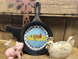 Decorative cast iron skillet with a cross stitched design on a wooden surface with farm-themed figurines.