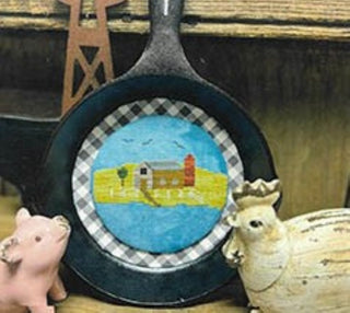 Decorative cast iron skillet with a cross stitched scenic design on a checkered background, surrounded by small figurines.