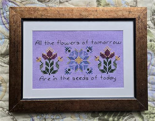 Framed cross-stitched artwork with flowers and text on a textured fabric background