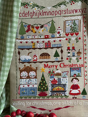 Cross stitch sampler featuring alphabet letters, holiday decorations, a Christmas tree, fireplace, and woman in red dress with stove — Waiting for Christmas One Stitch at a Time by Lilli Violette.