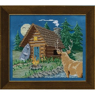 Cross-stitched scene of a cabin in the woods with a deer in the foreground, framed in brown.