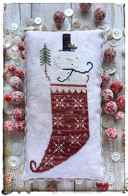 Cross-stitched red stocking with white snowflake motifs and a snowman peeking out of the top, holding a tiny evergreen tree, finished as a small pillow and displayed with frosted red berries and white buttons.